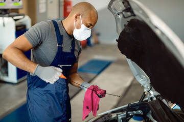 Black auto mechanic checking car oil while wearing face mask in repair shop.