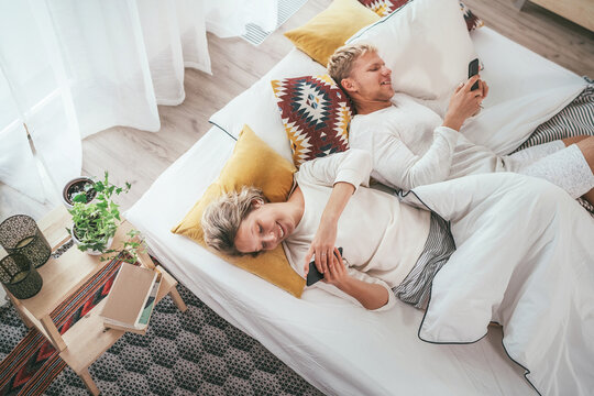 Top view of the couple lying in cozy bed in bedroom and browsing internet and checking mails using each other modern smartphone. Modern technology, social issues and relations problems concept image.