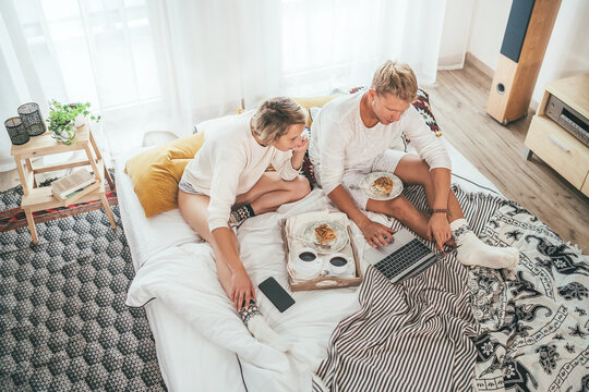 Top view of the couple in pajamas in cozy bed browsing internet together using a laptop and having a morning coffee with apple pie dessert. . Couples relations and internet technology concept image.