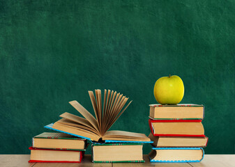 Open textbook, pile of books in colorful covers and green apple on wooden table with green blackboard background. Distance home education. Back to school, quarantine concept of stay home.