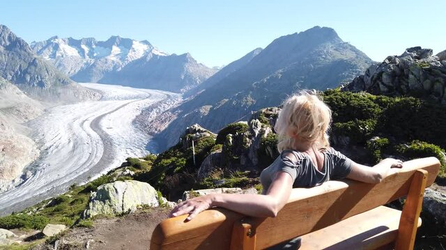 Aletsch Glacier from Moosfluh viewpoint in summer with clear blue sky in Swiss Alps, Valais Canton, Switzerland, Europe. Tourist woman relaxing sitting in a wooden bench looking over glacier.