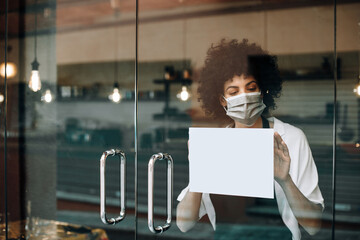 Restaurant female owner with protective face mask, reopens after lockdown quarantine. Business woman hangs a blank paper on a glass door. Mockup for your idea. New rules during Coronavirus pandemic