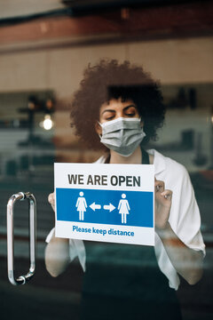 Coffee Shop Female Owner With Protective Face Mask, Reopens After Lockdown Quarantine. Business Woman Hangs A Open Sign That Asks To Keep Distance. New Rules During Coronavirus Pandemic