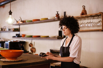 Portrait of a caucasian female barista working with the card terminal. Woman with apron standing behind the bar counter of a restaurant. Small business owner, young entrepreneur conceptual.