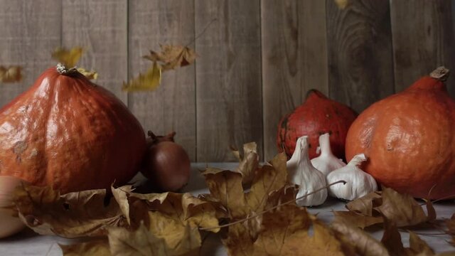 Dry leaves are falling at the wooden table with pumpkins and garlic in slow motion. Leaf foliage is moving at the table with still life of fall symbols. Theme of thanksgiving and autumn vegetables.