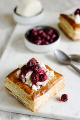 Puff pastry squares with ricotta and cherry sauce on white marble tray. Baked small square cakes topped with cheese and berry sauce