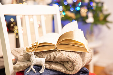 Christmas hygge background with warm sweaters, book, christmas decoration toy on festive bokeh background. Reading, cozy home, holidays, xmas concept