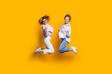 Fototapeta na wymiar Two blonde sisters jumping on a yellow studio wall and smile at camera gesturing happiness