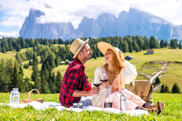 Young couple in love doing picnic visiting alps dolomities. Boyfriend and girlfriend laughing together drinking red wine sitting on green beautiful field.