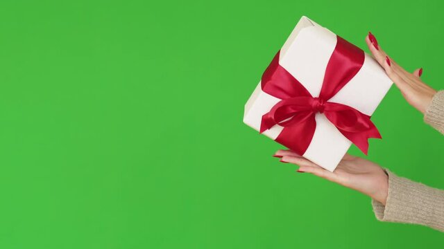 Holiday present. Christmas gift delivery. Festive surprise. Female hands showing white wrapped box with red ribbon bow isolated on green empty space background.