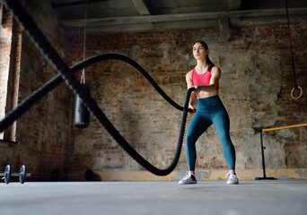 Young woman exercising with battle ropes at the gym. Strong female athlete doing crossfit workout...