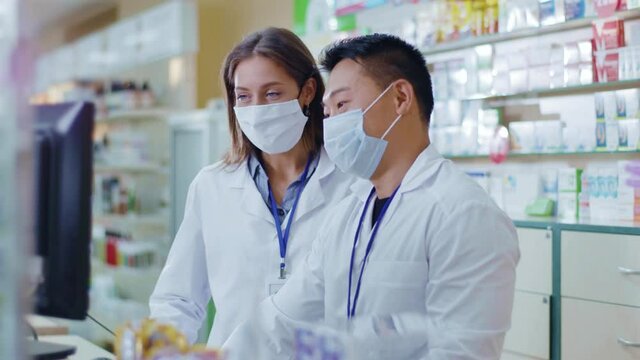Mixed-race couple asian male and caucasian female pharmacists in face masks examining drugs use using computer database. Collaboration. Pharmacy. Medicare.