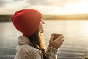 Woman in a red hat is drinking coffee on the lake at sunset. Autumn outdoor recreation. Concept of freedom of travel