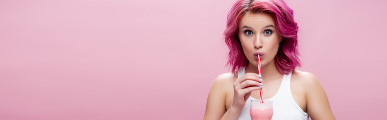 surprised young woman with colorful hair drinking strawberry milkshake from straw isolated on pink, panoramic shot