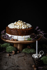 Homemade Carrot cake with ricotta cheese and honey frosting on wooden cake stand surrounded with moss, acorns, pines and with little white candle on foreground on black background.