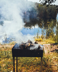 Metal brazier with fire wood and big smoke close up at nature background, near lake with trees and green grass, grill barbeque, picnic, fishing summer weekend concept outdoors 