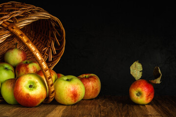 apples scattered from a wicker basket lying on its side on a wooden table surface against a black concrete wall. dark artistic shot with copy space. autumn fruit concept