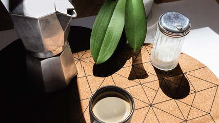 Comienza el día con un delicioso café. Vista cenital de una taza de café bajo el sol junto a una antigua cafetera italiana y un azucarero sobre un mantel de corcho con estilo y formas geométricas.