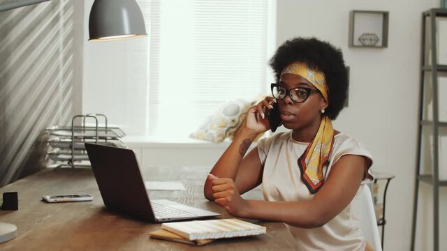 Young beautiful Afro-American businesswoman in casualwear sitting at desk in office and speaking on mobile phone