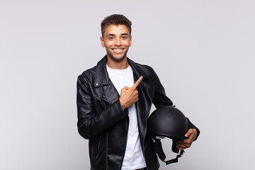 young motorbike rider smiling cheerfully, feeling happy and pointing to the side and upwards,...