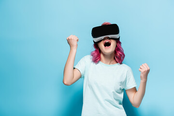 excited young woman with pink hair in vr headset showing yes gesture on blue background