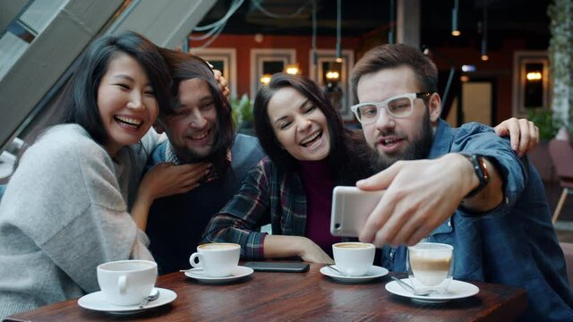 Multi-ethnic group of friends Asian and Caucasian are taking selfie in cafe using smartphone camera posing with funny faces and hair. Youth lifestyle concept.