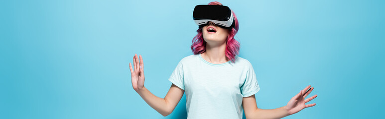 shocked young woman with pink hair in vr headset gesturing on blue background, panoramic shot