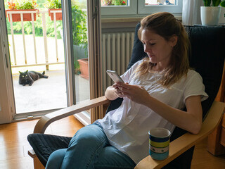 Young woman uses smartphone while her cat watches her on the terrace, cat owner at home while using...