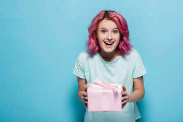 excited young woman with pink hair holding gift box with bow on blue background