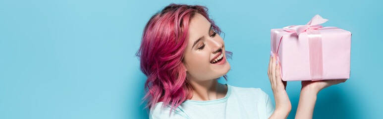 Fototapeta premium young woman with pink hair holding gift box with bow and smiling on blue background, panoramic shot