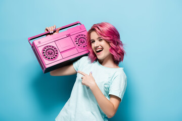 excited young woman with pink hair pointing at vintage tape recorder on blue background