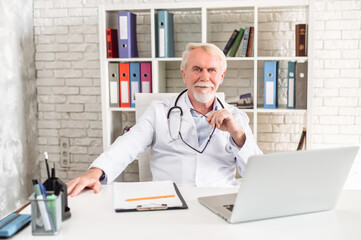 Mature male gray-haired doctor sits at the desk with a laptop and looks at camera with a pleasure...