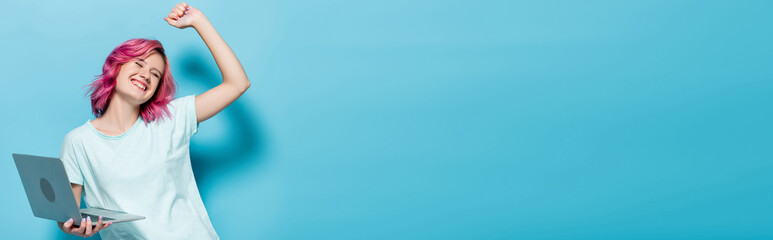 young woman with pink hair holding laptop and smiling on blue background, panoramic shot