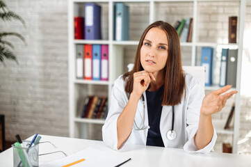 Webcam view of young puzzled female doctor in white wear with a stethoscope on the shoulders, she looks at camera and listens with a serious face. Online visit concept