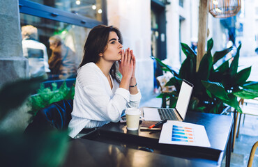 Contemplative female freelancer pondering on diagram statistics editing using blank laptop computer with copy space, thoughtful woman with netbook thinking during distance paperwork at street terrace