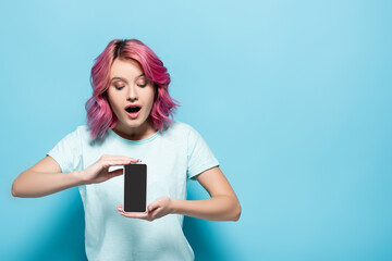 Naklejka premium shocked young woman with pink hair holding smartphone with blank screen on blue background
