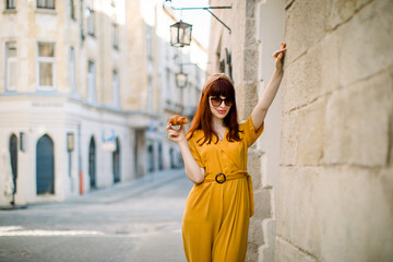 Fototapeta na wymiar Fashion city portrait of gorgeous young Caucasian red haired woman, dressed in stylish yellow overalls, sunglasses and head hoop, walking in old European city with fresh tasty croissant