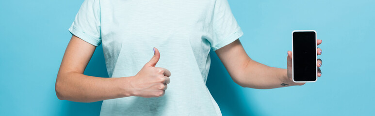 cropped view of young woman holding smartphone with blank screen and showing thumb up on blue background, panoramic shot