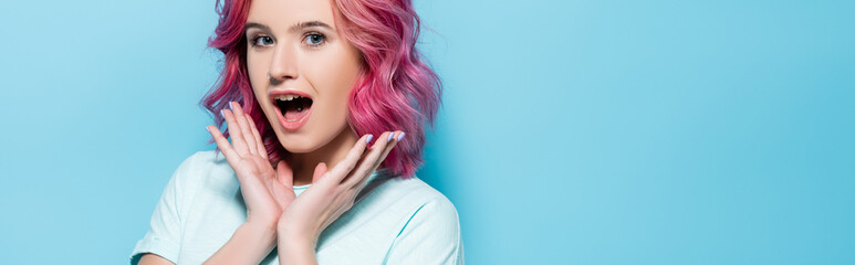 surprised young woman with pink hair and hands near face on blue background, panoramic shot