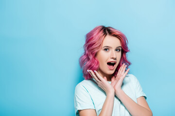 surprised young woman with pink hair and hands near face on blue background