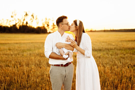 Beautiful family with little daughter walking at the field, happy parents hold cute newborn baby girl in arms, handsome father kiss gorgeous mother, parenthood concept