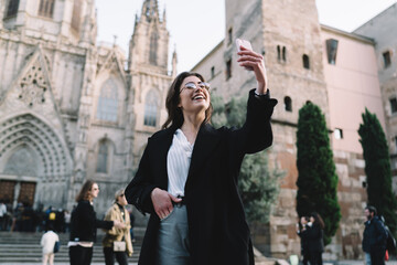 Happy female blogger shooting influence vlog for making online sightseeing at Italian piazza with ancient duomo, cheerful hipster girl in glasses smiling at camera while making front video translation