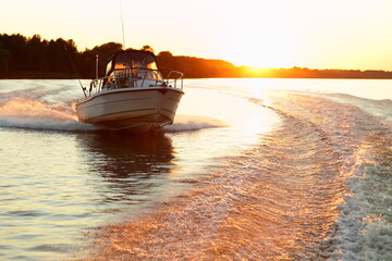 Fast gliding motor boat in wake track on beautiful sunset background, bright orange sun shine in calm water on forest stripe on Horizon at Sunny summer evening, front side view from powerboat stern - obrazy, fototapety, plakaty