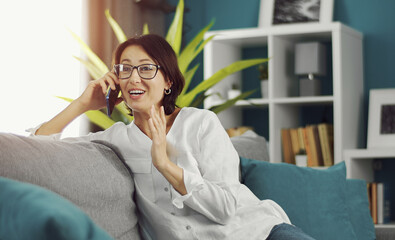 Portrait of cheerful adult woman talking on smartphone sitting sideways on couch in flat