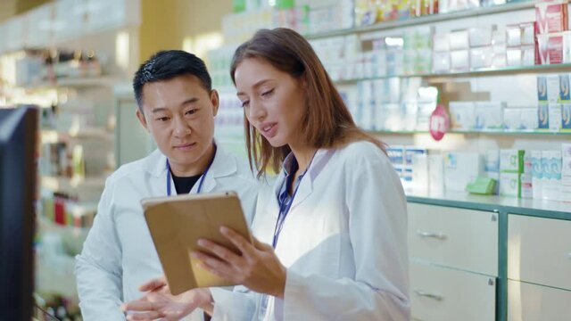 Asian adult and younger female intern cooperating in drugstore. Aspiring woman apothecarist asking advice from male colleague learning drugs in pharmacy using tablet.