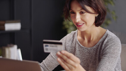 Cheery woman holding credit card paying online for purchase using laptop, focus on face