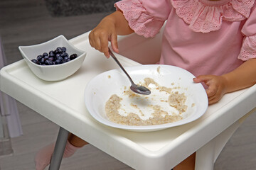 Little kid girl eating her morning porridge oatmeal with blueberries herself, sitting in baby chair in kitchen