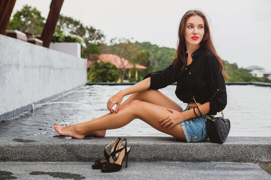 young sexy beautiful woman sitting at pool barefoot, long skinny tanned legs, high heeled shoes, black shirt, denim shorts, purse, relaxing, summer style, vacation, close-up details