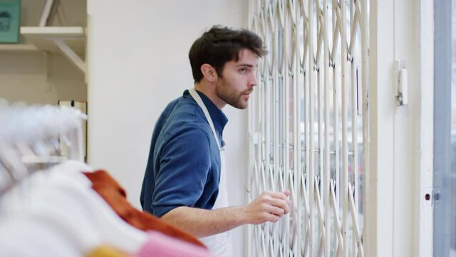 Man running small business pushing back security grill from window and opening shop viewed from inside - shot in slow motion