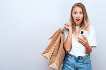 Young female in casual cloth with paper shopper bags holding and looking to smartphone with shocked...